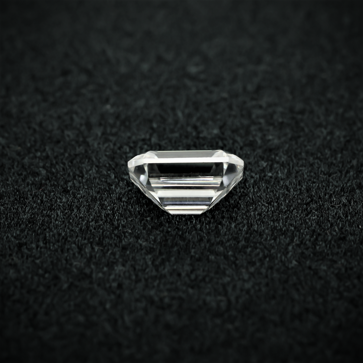 Back view of an emerald cut moissanite resting on a black textured surface, showcasing the culet and step cut pavilion facets with sharp clarity